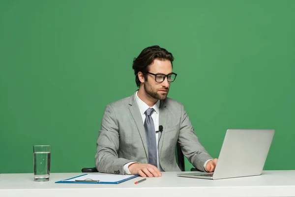 Presentador de noticias barbudo en gafas y traje usando portátil aislado en verde - foto de stock