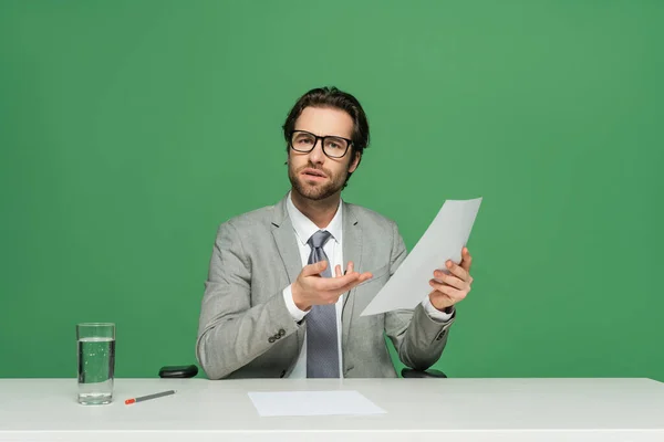 Confuso presentador de noticias en anteojos y traje apuntando con la mano a papel en blanco aislado en verde - foto de stock