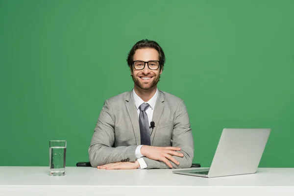 Presentador de noticias barbudo en gafas y traje sonriendo mientras está sentado cerca de la computadora portátil aislado en verde - foto de stock