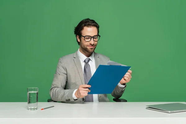 Sonriente presentador de noticias en gafas y traje sujetando portapapeles cerca del ordenador portátil aislado en verde - foto de stock