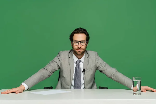 Broadcaster in eyeglasses and grey suit sitting at desk isolated on green — Stock Photo