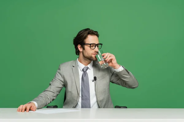Broadcaster in eyeglasses sitting at desk and drinking water isolated on green — Stock Photo