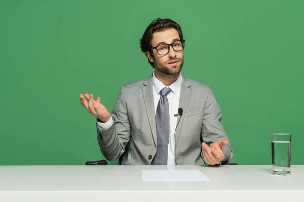 Locutor de noticias en gafas y traje sentado en el escritorio y hablando aislado en verde - foto de stock