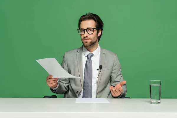 Locutor de noticias en gafas y traje sentado en el escritorio y sosteniendo papel aislado en verde - foto de stock