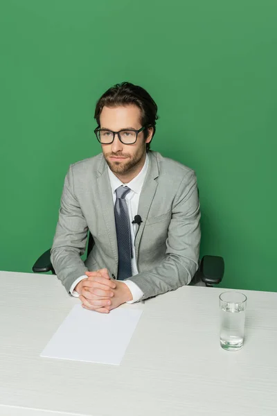 Vista aérea de emisora de noticias en gafas y traje sentado con las manos apretadas en el escritorio aislado en verde - foto de stock