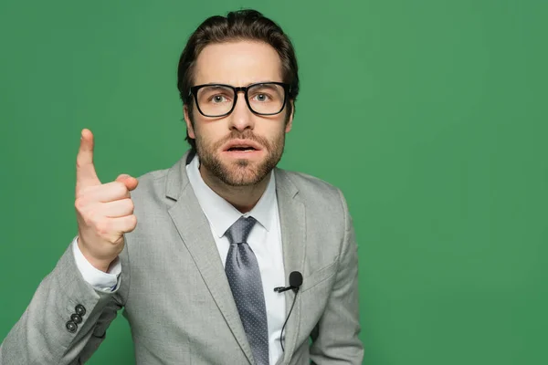 Emotional newscaster in eyeglasses and suit pointing with finger isolated on green — Stock Photo