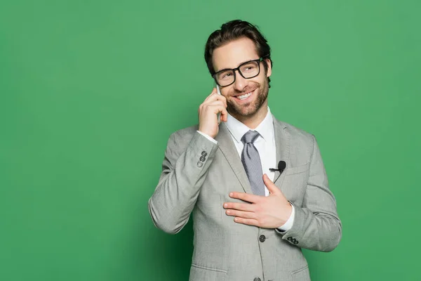 Fröhlicher Reporter in Brille und Anzug, der auf grünem Hintergrund mit dem Smartphone spricht — Stockfoto