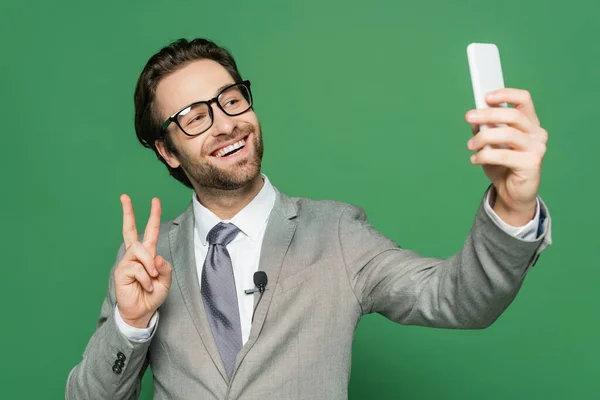 Reportero sonriente en anteojos y traje tomando selfie en smartphone aislado en verde - foto de stock