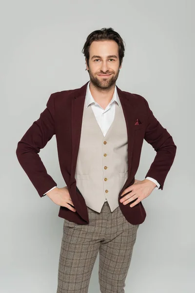 Cheerful showman in vest and red blazer standing with hands on hips isolated on grey — Stock Photo
