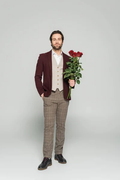 Full length of host of event in blazer holding red roses and standing with hand in pocket isolated on grey — Stock Photo