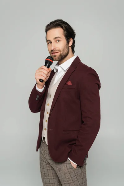 Bearded host of event in formal wear talking in microphone and standing with hand in pocket isolated on grey — Stock Photo