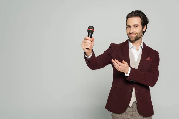 Cheerful host of event in formal wear pointing at microphone isolated on grey — Stock Photo