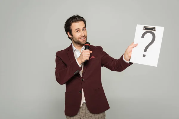 Bearded host of event in formal wear talking in microphone while holding clipboard with question mark isolated on grey — Stock Photo