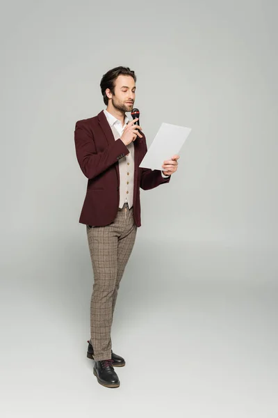 Full length of bearded showman in formal wear talking in microphone and looking at paper on grey — Stock Photo
