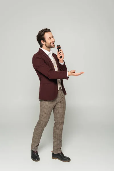 Full length of bearded showman in formal wear singing in microphone on grey — Stock Photo