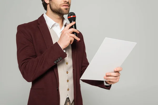 Vista ritagliata di showman barbuto in abbigliamento formale parlando in microfono e tenendo carta su grigio — Foto stock