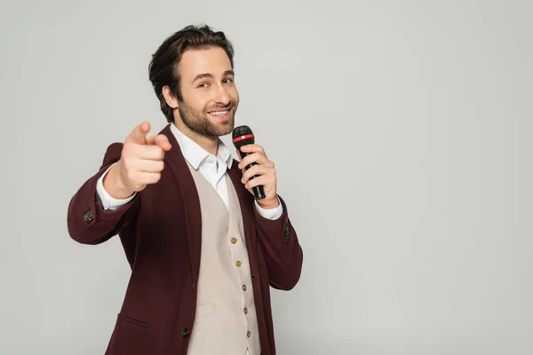 Felice showman in abito formale parlando in microfono e puntando con il dito alla fotocamera isolata su grigio — Foto stock