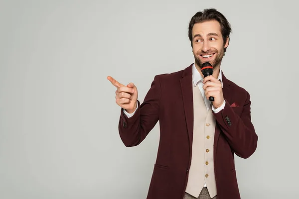Positive showman in formal wear talking in microphone and pointing with finger isolated on grey — Stock Photo