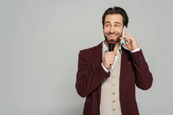 Щасливий шоумен говорить на смартфоні, тримаючи мікрофон ізольовано на сірому — стокове фото