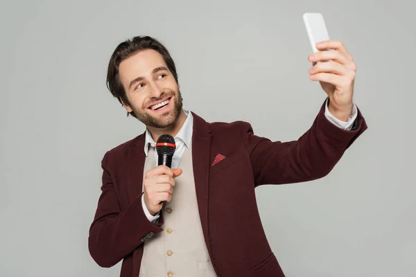 Cheerful showman taking selfie on smartphone while holding microphone isolated on grey — Stock Photo
