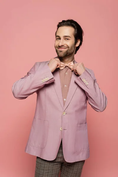 Cheerful and young showman in suit adjusting bow tie isolated on pink — Stock Photo