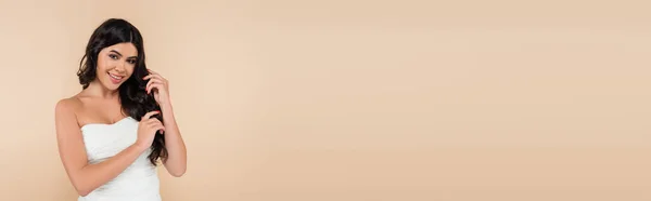 Positive brunette woman in top touching wavy hair isolated on beige, banner — Stock Photo