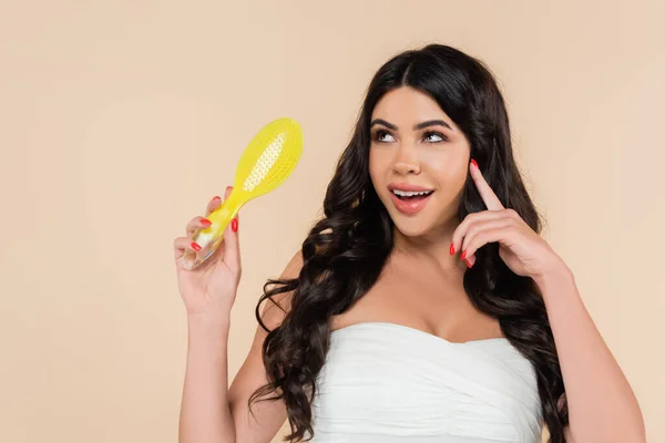 Dreamy woman in top holding hairbrush and looking away isolated on beige — Stock Photo