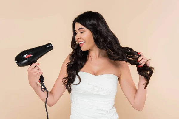 Cheerful brunette woman holding hairdryer and touching hair isolated on beige - foto de stock
