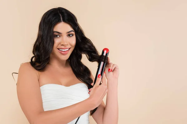 Cheerful brunette woman using curling iron isolated on beige with copy space — Stock Photo