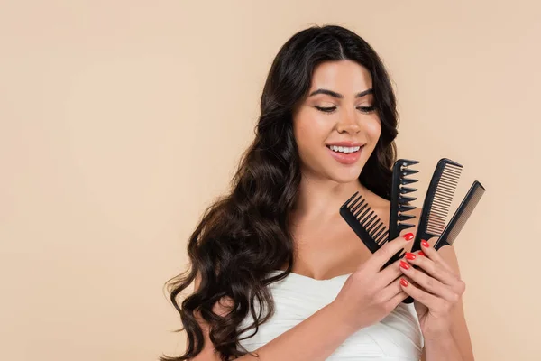 Brunette woman with wavy hairstyle looking at combs isolated on beige - foto de stock