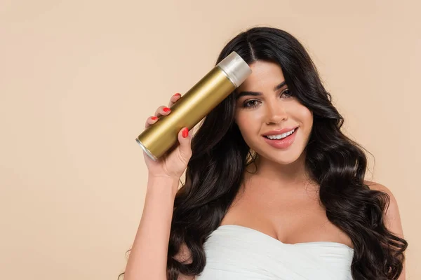 Positive brunette woman holding hairspray and looking at camera isolated on beige - foto de stock