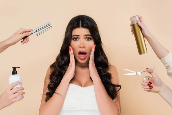 Shocked brunette woman looking at camera near hands with hairdressing tools isolated on beige — Stock Photo