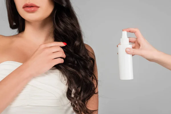 Cropped view of hair stylist holding spray bottle near brunette woman with shiny hair isolated on grey — Stock Photo