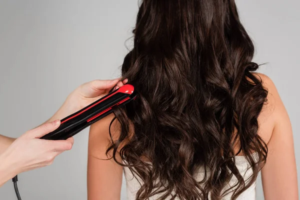 Vista ritagliata della parrucchiera che tiene la piastra dei capelli vicino alla donna bruna con i capelli ricci isolati sul grigio — Foto stock