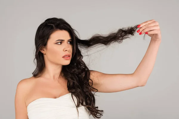 Donna scontenta in top bianco guardando i capelli danneggiati e aggrovigliati isolati su grigio — Foto stock