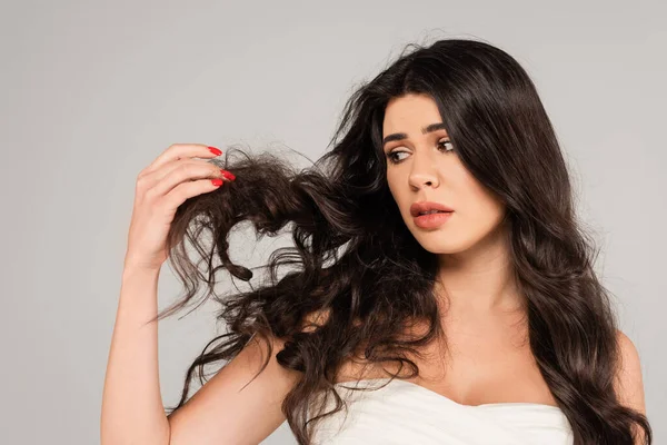 Displeased brunette woman looking at long and tousled hair isolated on grey — Stock Photo