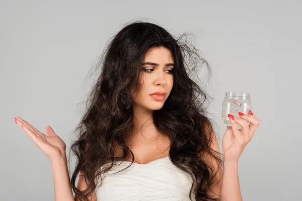 Brunette woman with tangled hair showing shrug gesture while holding styling gel isolated on grey — Stock Photo