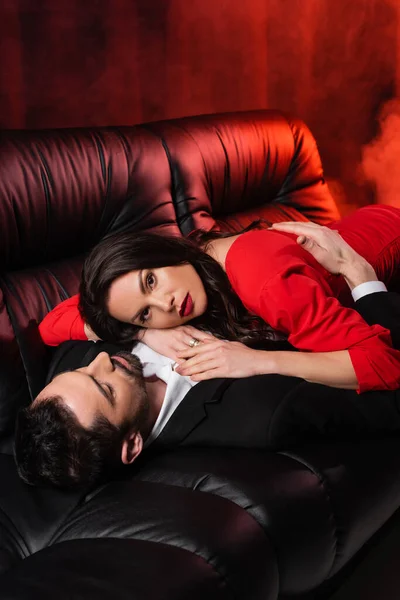 Brunette woman lying with bearded boyfriend on black couch on red with smoke - foto de stock