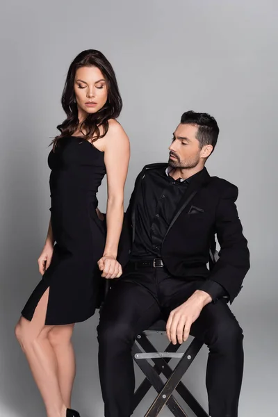 Bearded man sitting on chair while looking at sexy woman in black dress isolated on grey — Fotografia de Stock