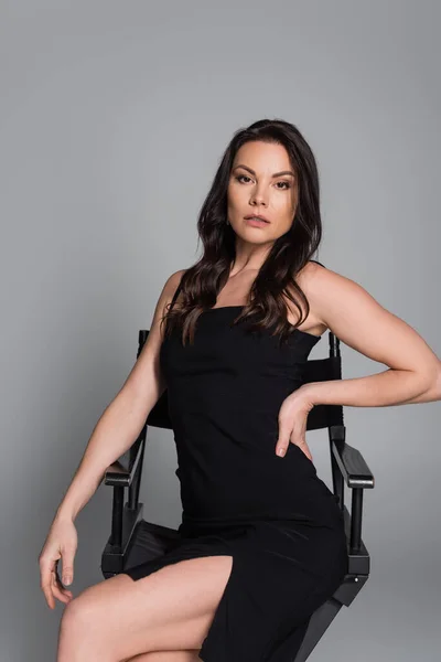 Brunette woman in black dress sitting on chair and posing with hand on hip on grey - foto de stock