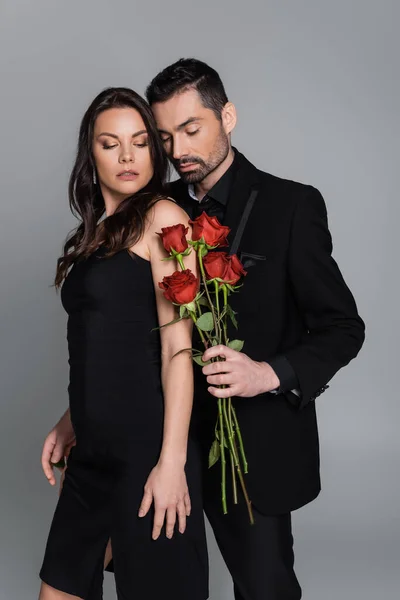Bearded man in suit holding red roses near brunette woman isolated on grey — Stock Photo