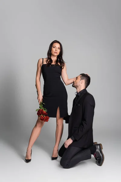 Full length of dominant woman in black dress holding red roses near bearded man in suit on grey — Stock Photo