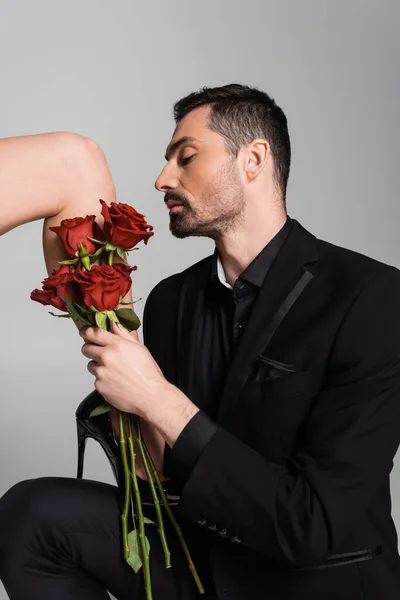 Submissive man in suit touching leg of dominant woman in high heeled shoe while holding roses isolated on grey — Stockfoto