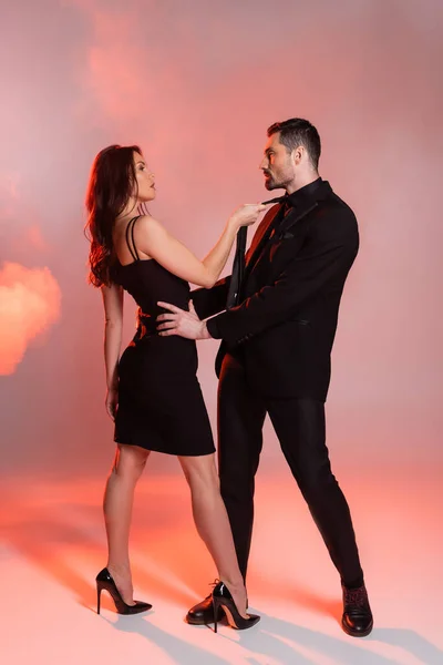 Full length of sexy woman in black dress pulling tie of man in formal wear on pink — Stock Photo