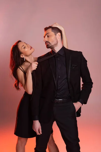 Sexy woman in black dress pulling tie while seducing confident man in formal wear on pink and grey — Stock Photo