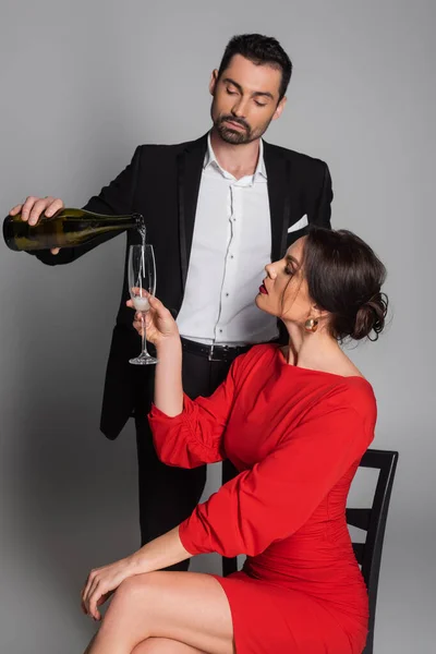Bearded man pouring champagne near girlfriend in red dress on grey background — Stock Photo