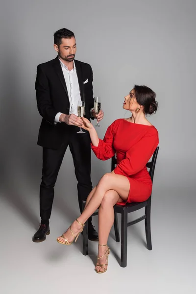 Full length of elegant man in suit giving glass of champagne to sexy girlfriend on grey background — Stock Photo