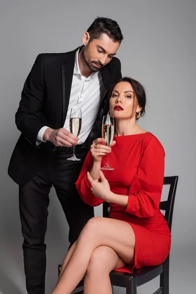 Elegant man in suit holding champagne near sexy girlfriend in dress sitting on chair on grey background — Stockfoto