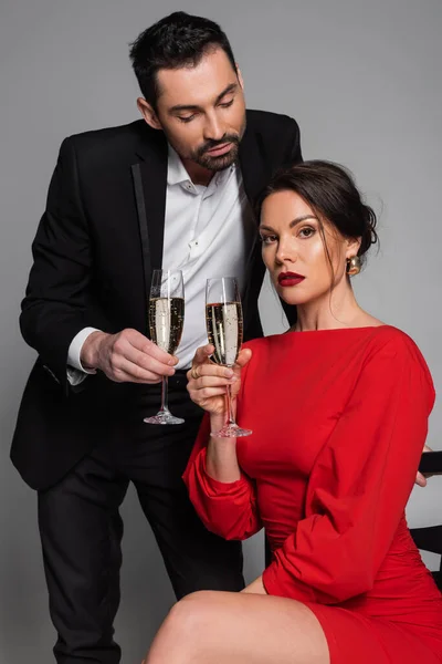Elegant man in suit holding champagne while girlfriend in dress looking at camera on grey background — Stock Photo