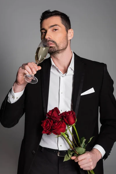 Elegant man in suit drinking champagne and holding red roses isolated on grey — Stock Photo